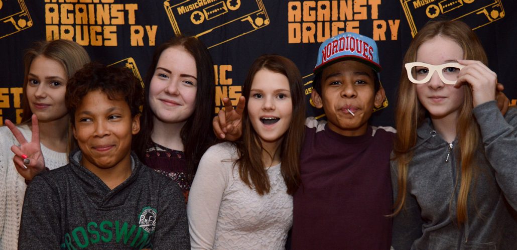 Kuusi yläkouluikäistä nuorta seisoo vierekkäin ja hymyilee Music Against Drugs -iltatapahtumassa.